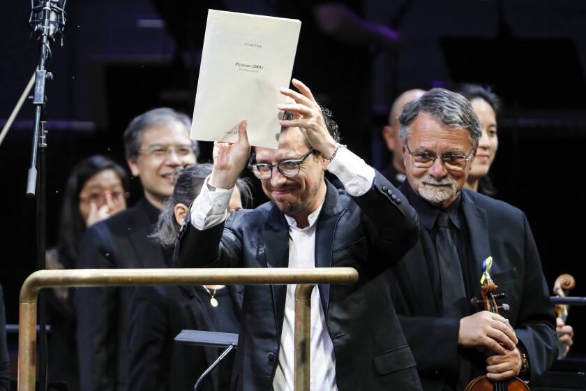 A man holds up paper, smiling. Behind him are orchestral musicians, standing and smiling.
