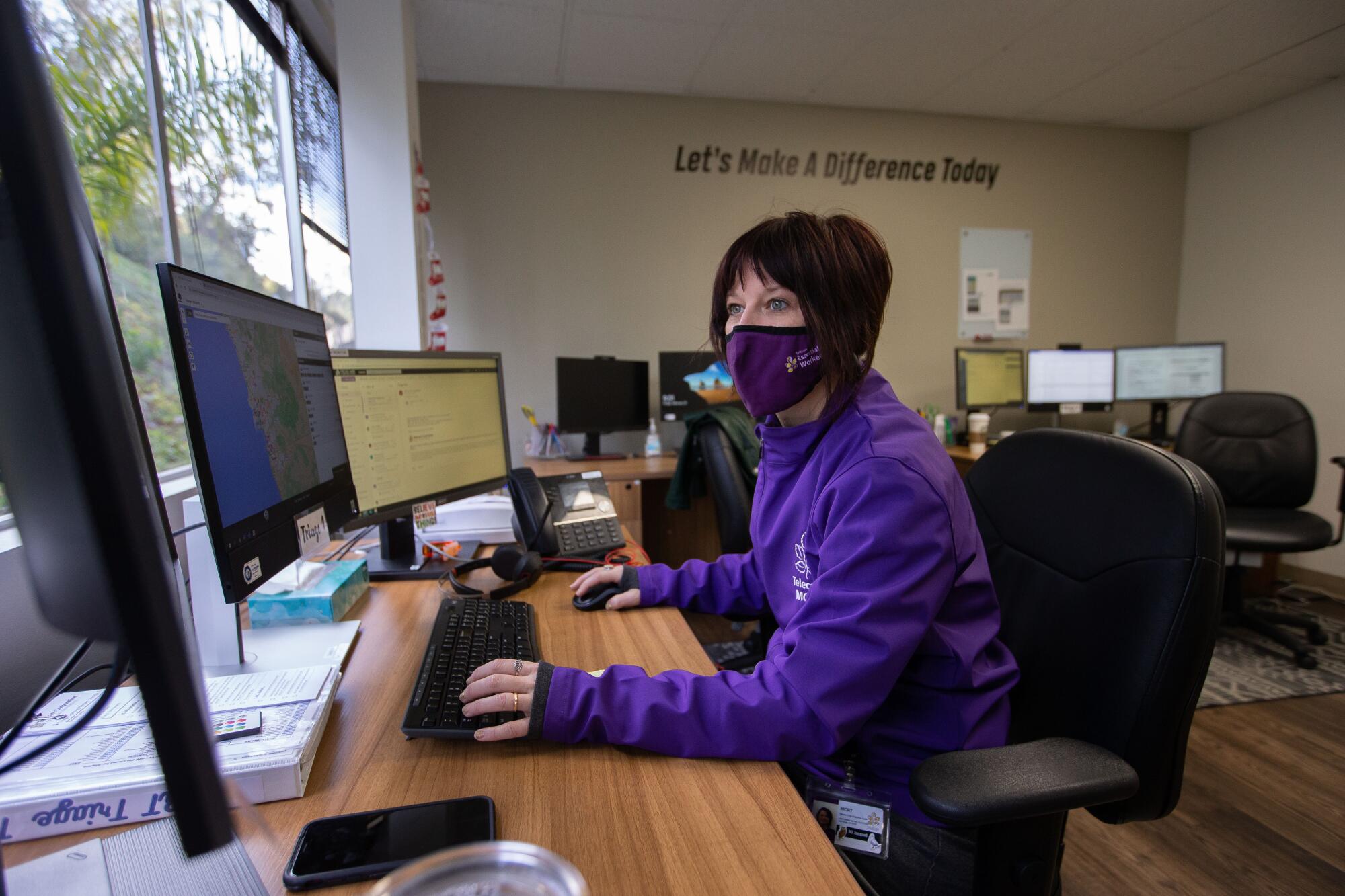 A woman wearing a purple mask and a purple shirt sits at a desk in front of a computer.