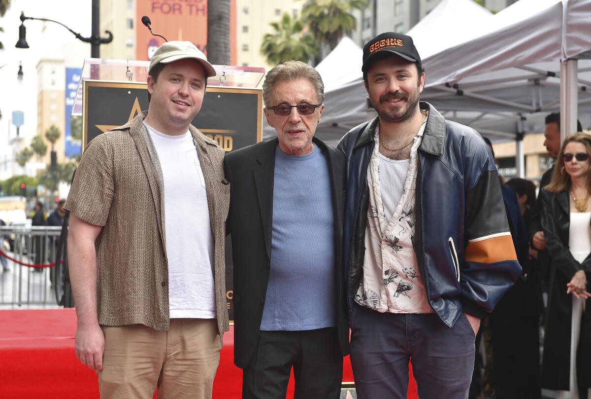 Frankie Valli wearing a blue shirt, a dark suit jacket, pants and sunglasses standing between two younger men in casual wear