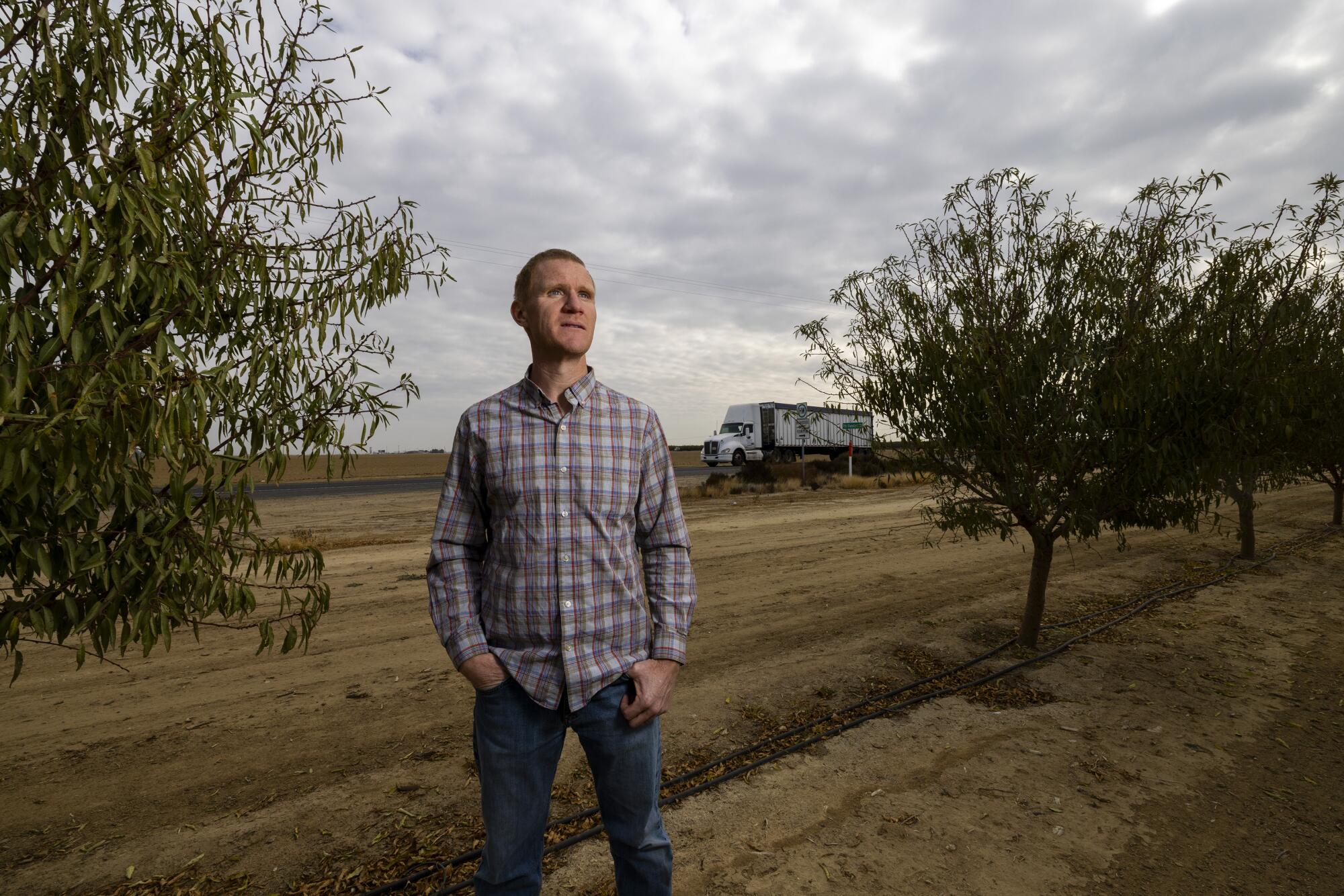 Daniel Palla stands among trees on farmland as a truck passes in the background.