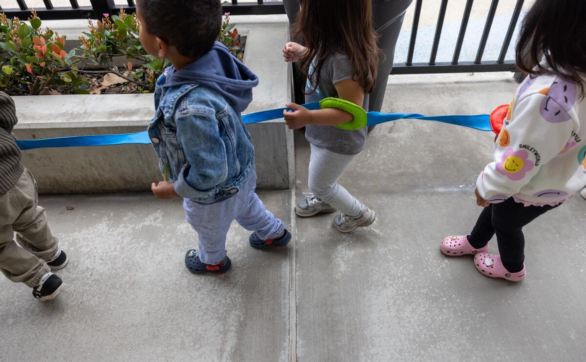 Toddlers walk connected by a cord near a fence 