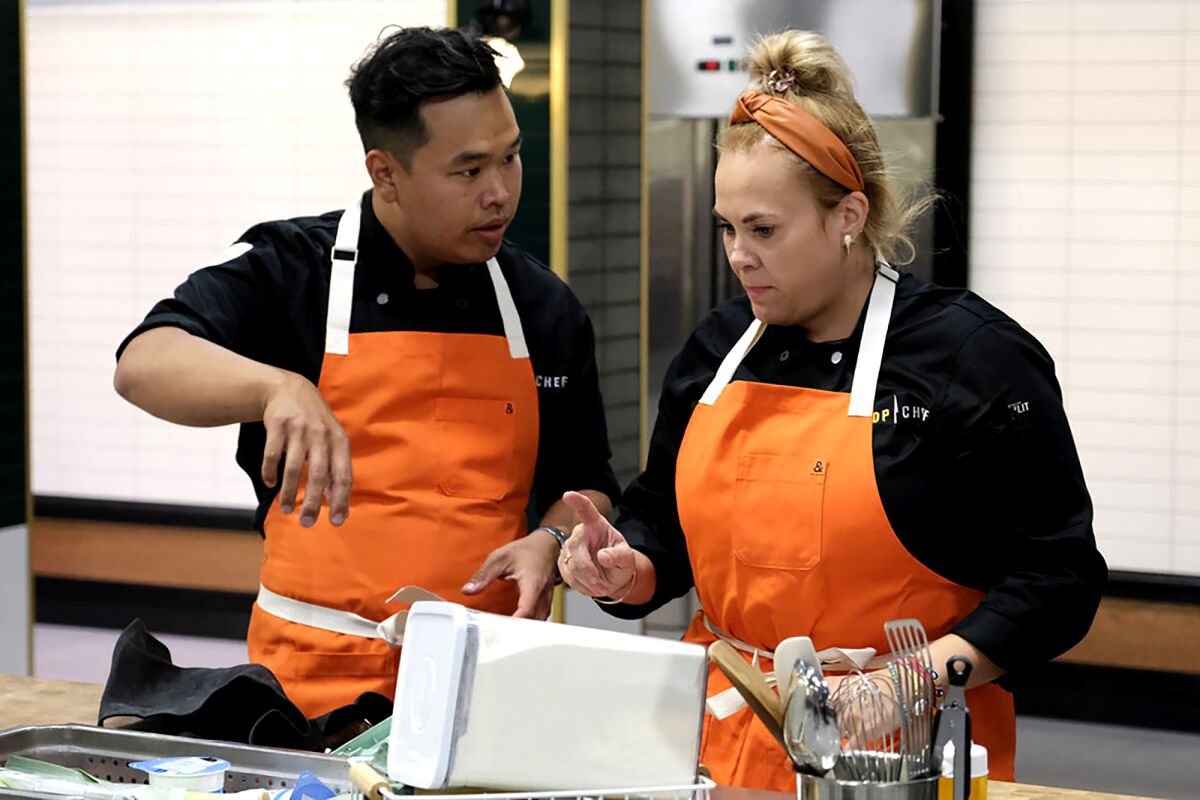 Buddha Lo and Sylwia Stachyra, who are both wearing black shirts and orange aprons, look at their kitchen tools on a table.