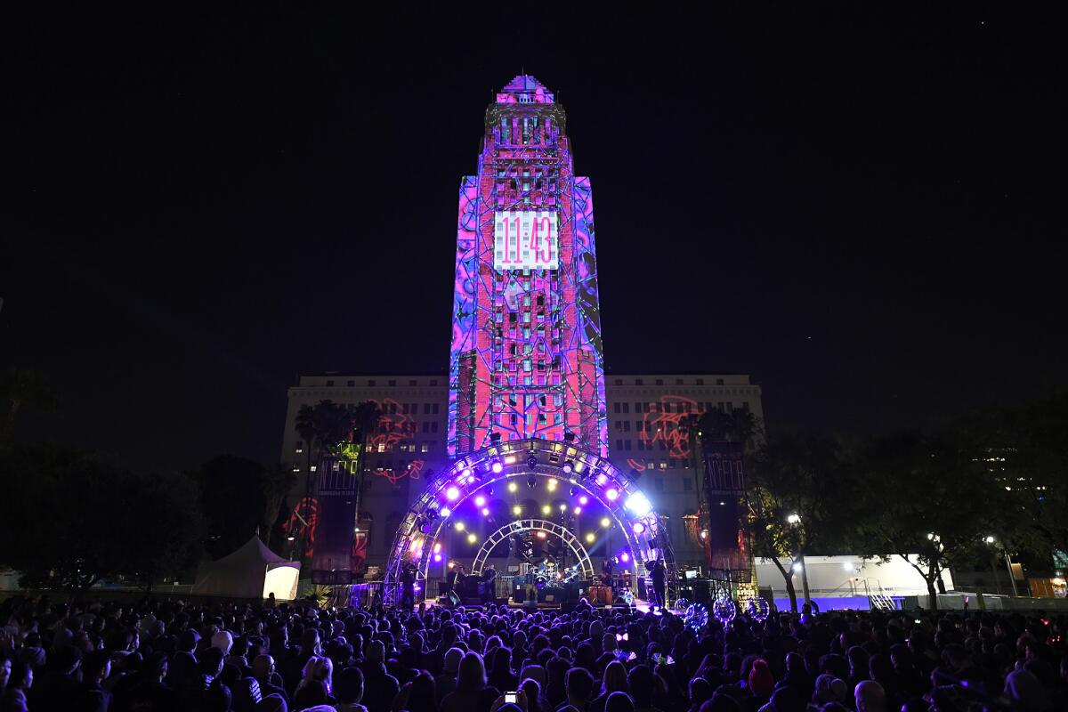 L.A. City Hall is seen illuminated with pink and purple lights and a projection of a countdown clock at 11:43 PM.