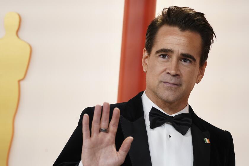 Colin Farrell in a tuxedo posing at the Oscars red carpet and waving his right hand