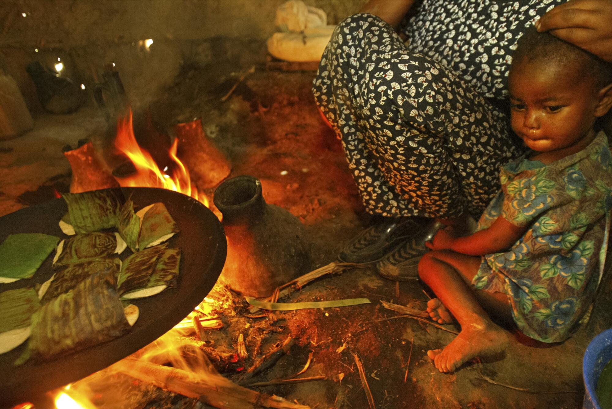 An infant sits with an adult near a cooking fire