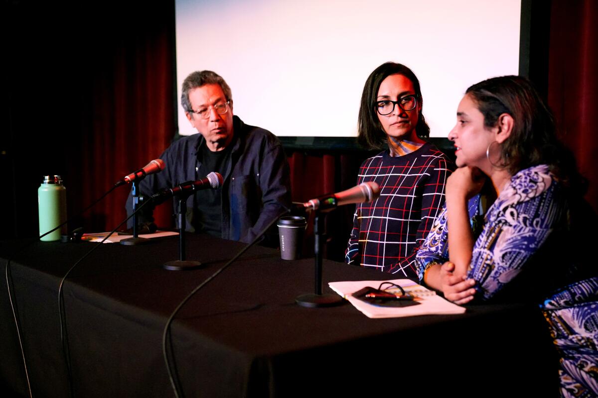 A man and two women share a table while speaking at mics.