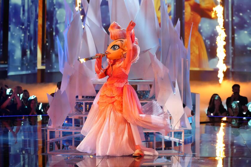 A person in an orange, goldfish-inspired gown with a goldfish headpiece performing on a stage 
