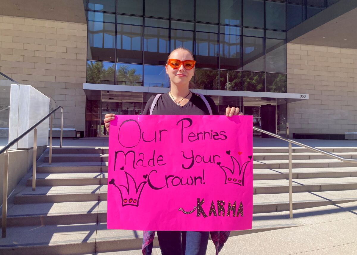 A Taylor Swift fan holds a pink paper sign that says, 'Our Pennies Made Your Crown. Karma'