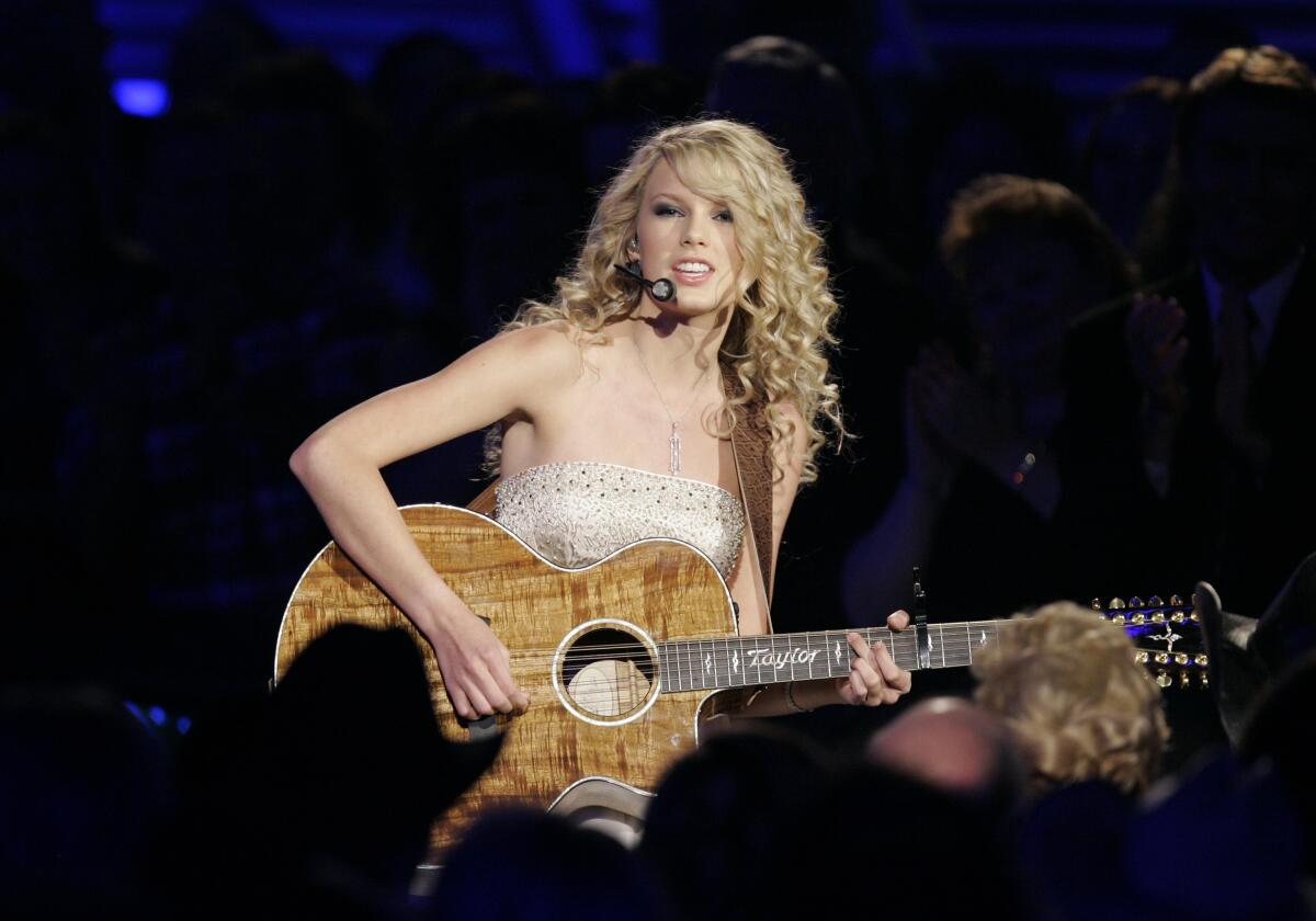 Taylor Swift, in a strapless top and with long curly hair, sits playing an acoustic guitar on a darkened stage