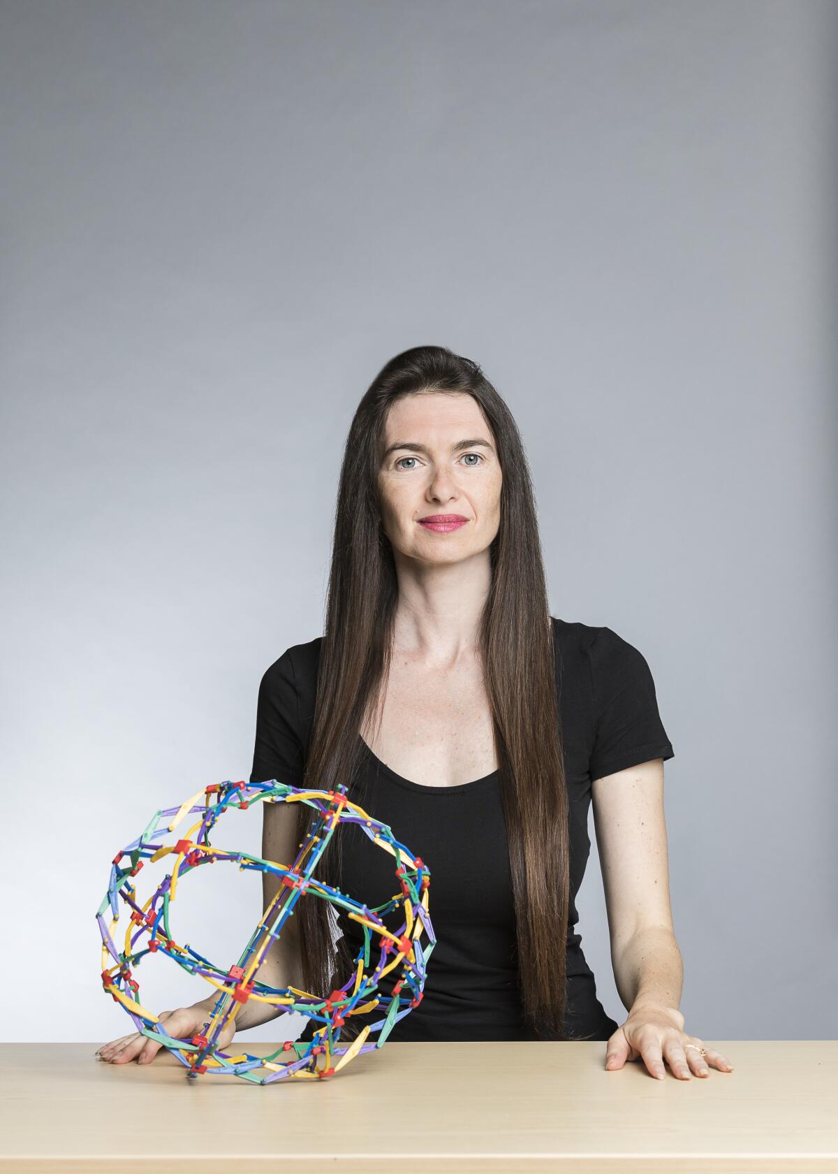 A woman in a black T-shirt stands behind a table with an expandable ball toy on it.