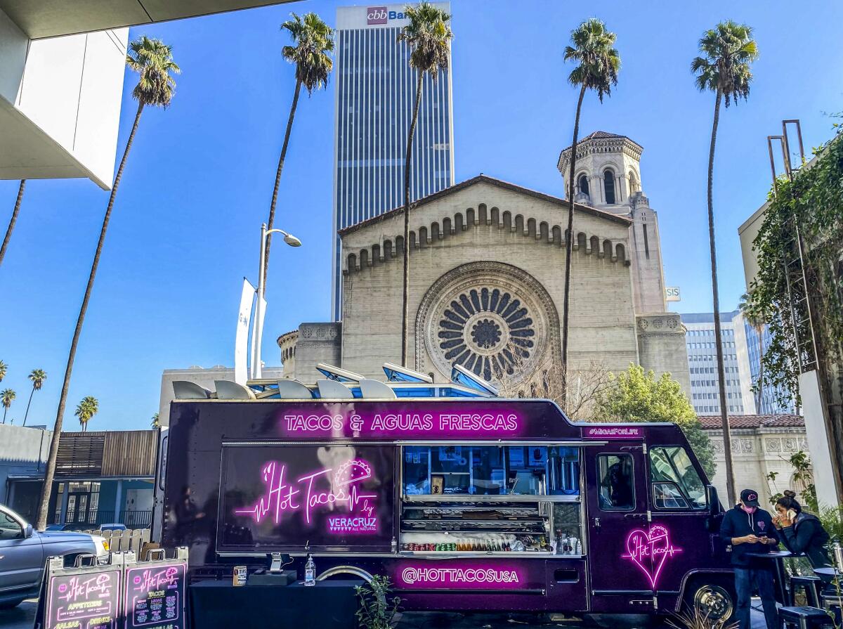 A black food truck on a city street has 'Hot Tacos' in pink neon on its side.