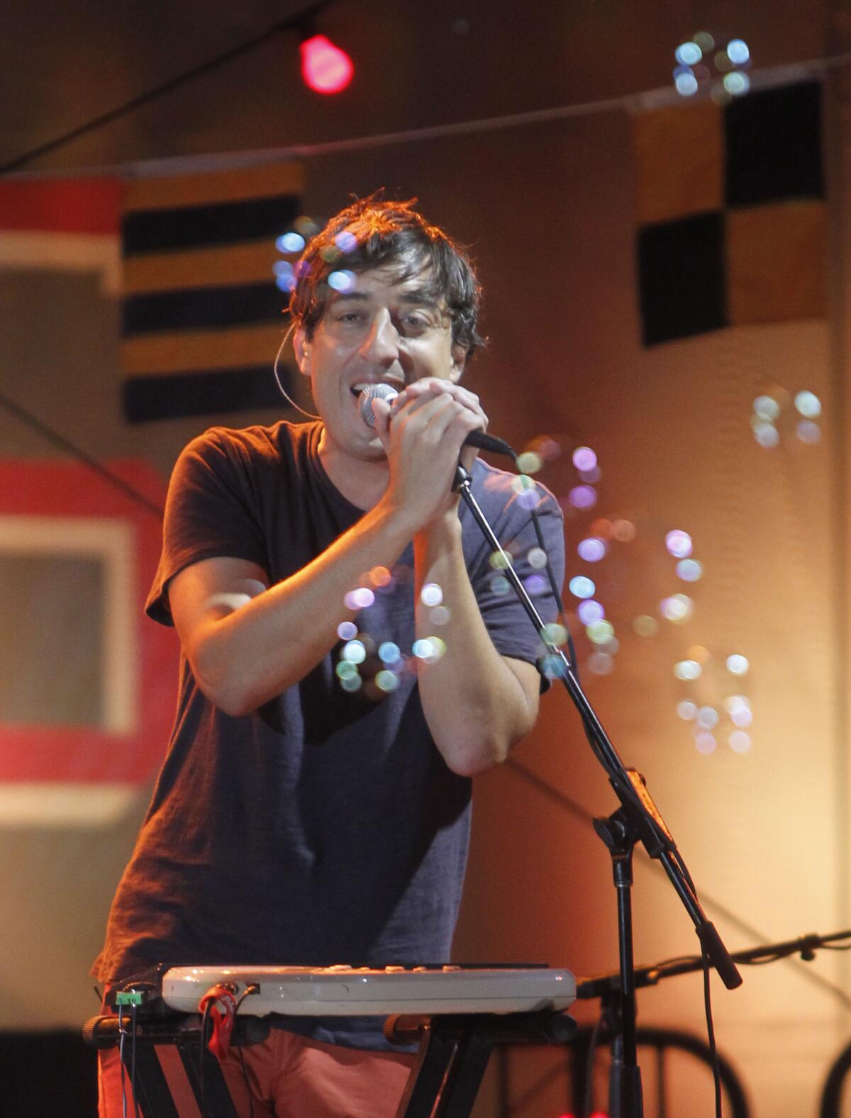 Ed Droste sings into a microphone behind a window in a recording studio.