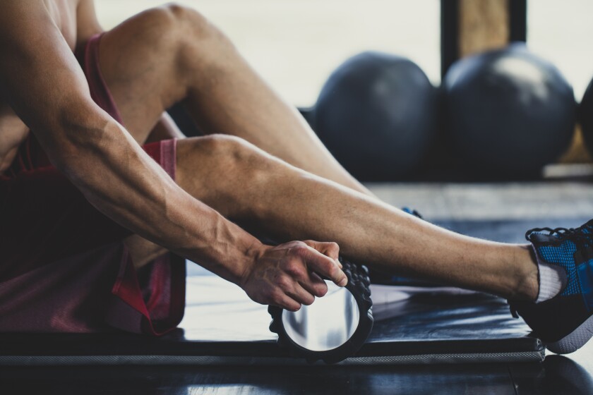 A muscular man massages his calves with a foam roller in an indoor gym.