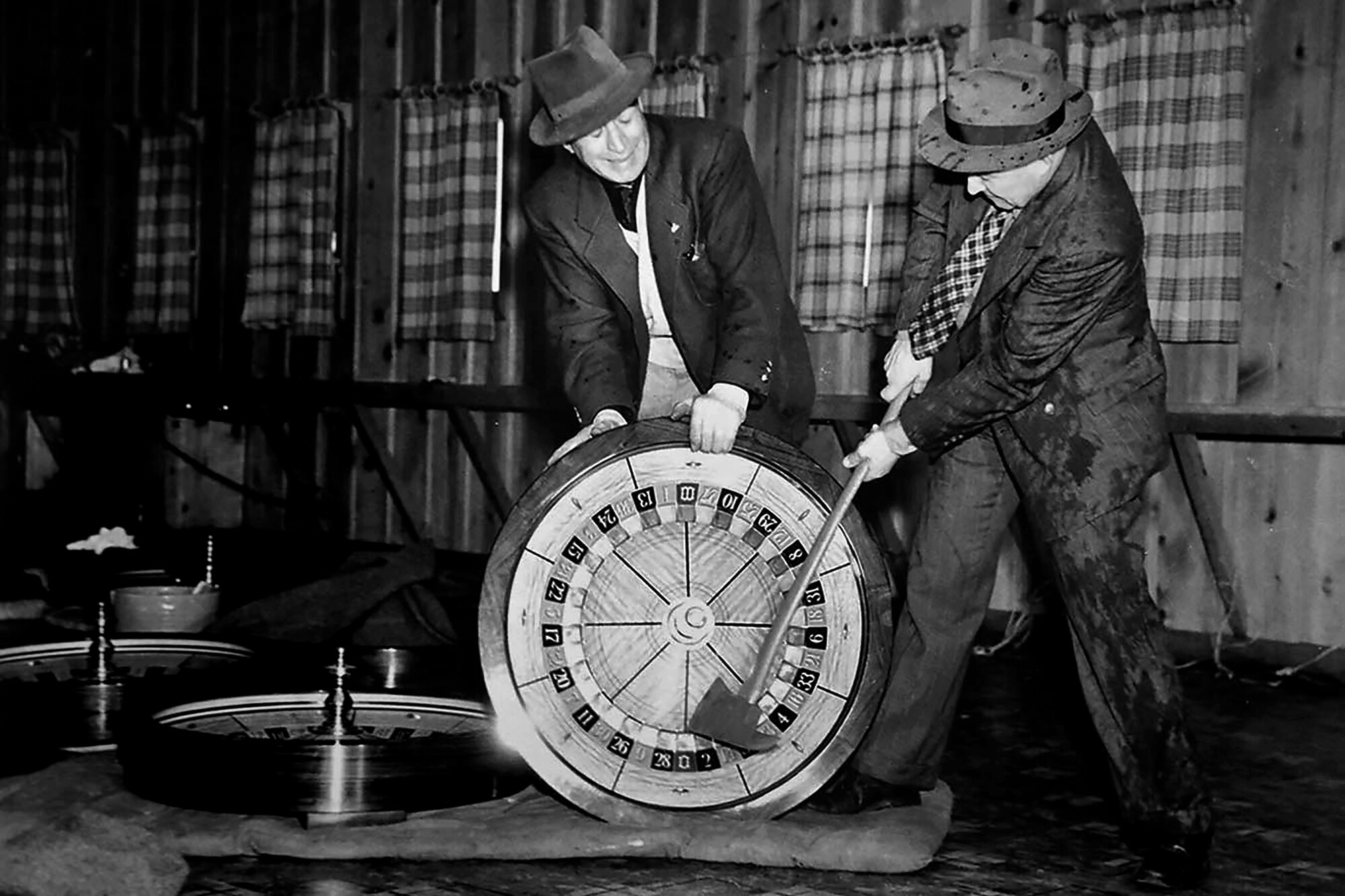An agent in a suit and hat takes an ax to a roulette wheel held by another agent