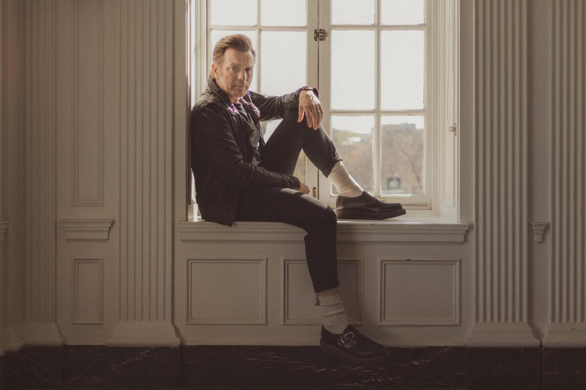 Ewan McGregor sits on a window seat with one knee raised.