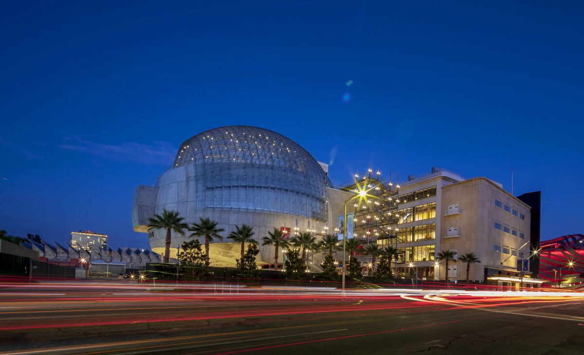 A large museum with a domed portion and palm trees in front is shown lighted up at dusk.