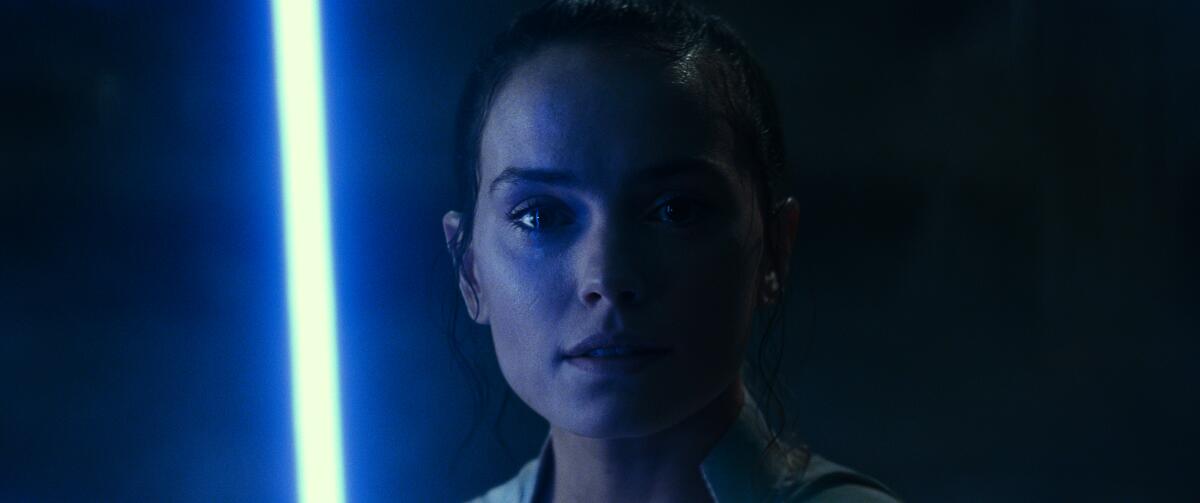 A woman stands in darkness with a glowing blue lightsaber blade to her right