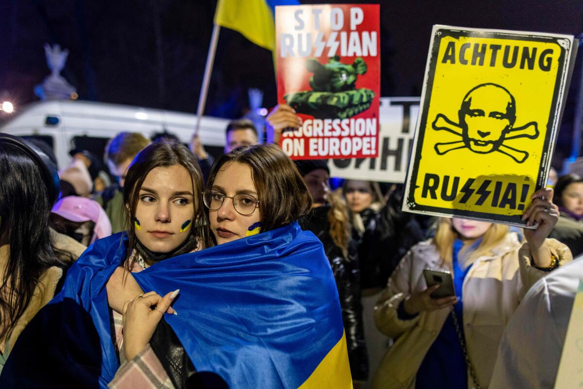 Two women with a yellow-and-blue flag around their shoulders and people holding up anti-Russia signs 