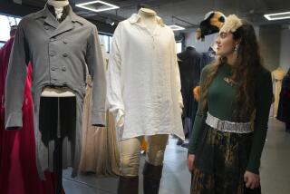 A woman with long brown hair in a green shirt and long skirt looking at a mannequin wearing a flowy white shirt and pants