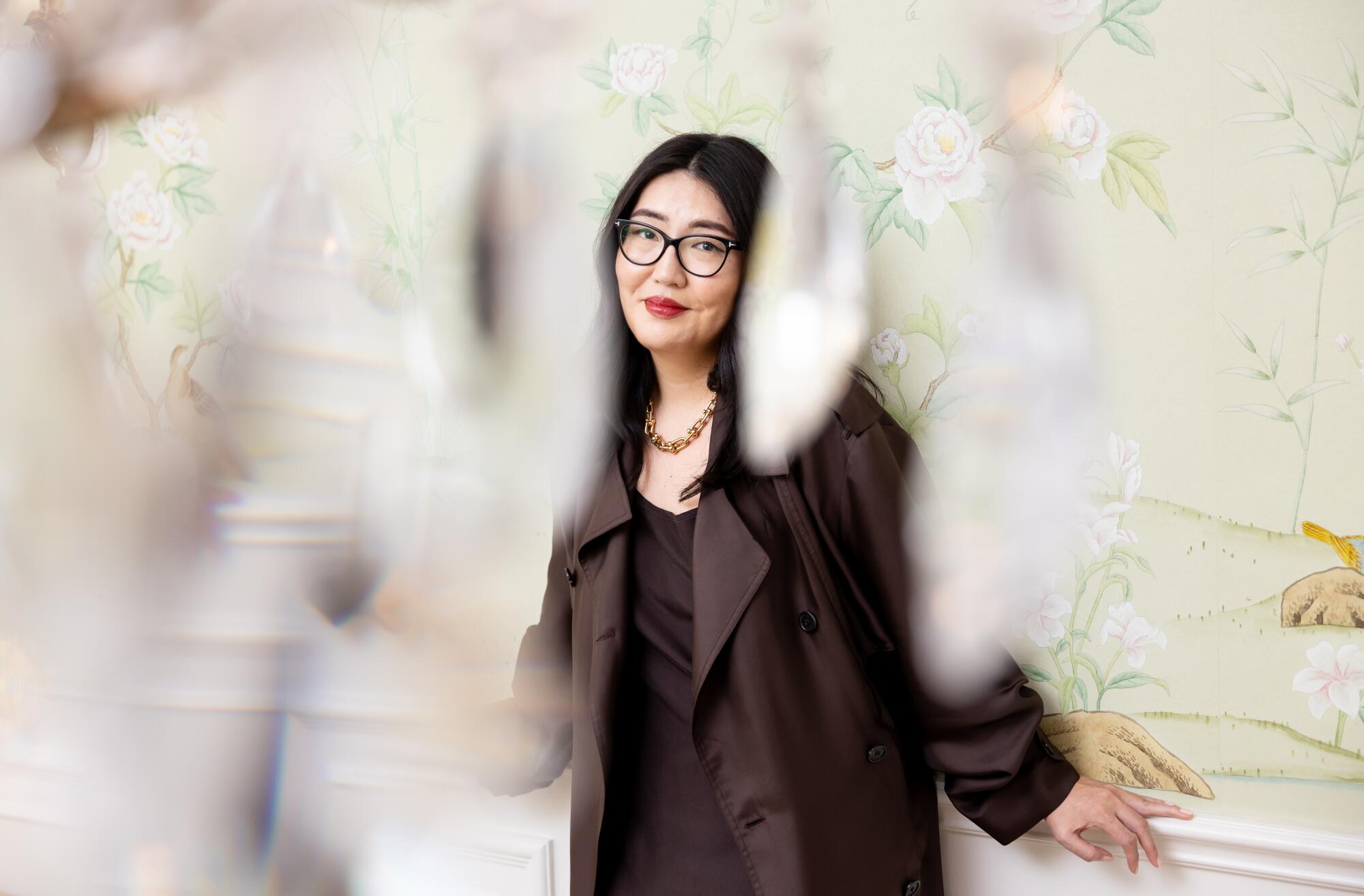 A woman with glasses in a black dress and coat, seen between the crystals of a chandelier, leans against a green floral wall.