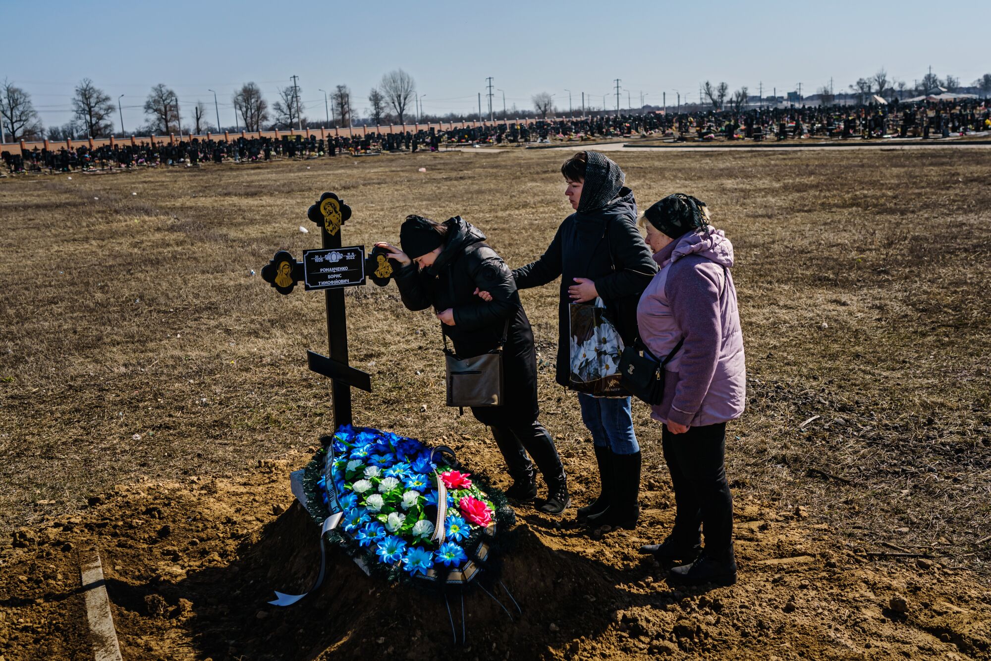A woman leans on cross marking a grave covered in blue, white and red flowers, as another supports her and a third stands by.