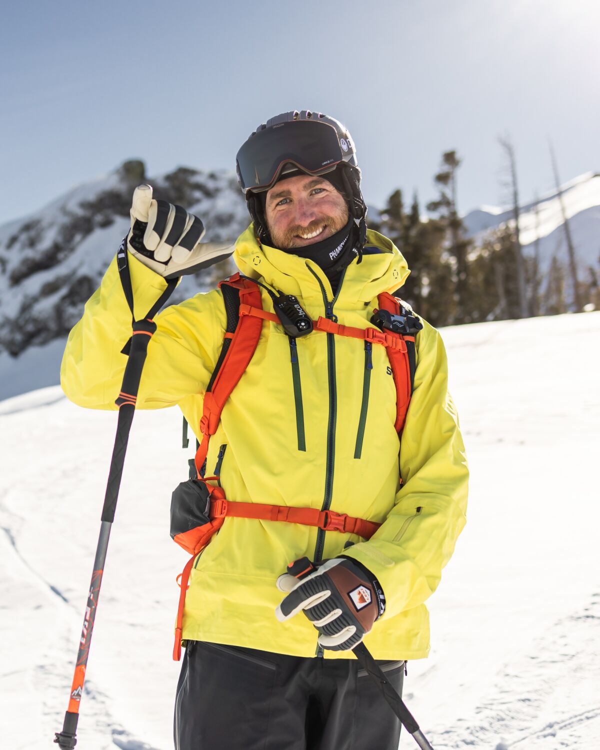 Vertical outdoors image of a smiling man with facial hair in skiing helmet, bright yellow jacket and holding skiing poles.