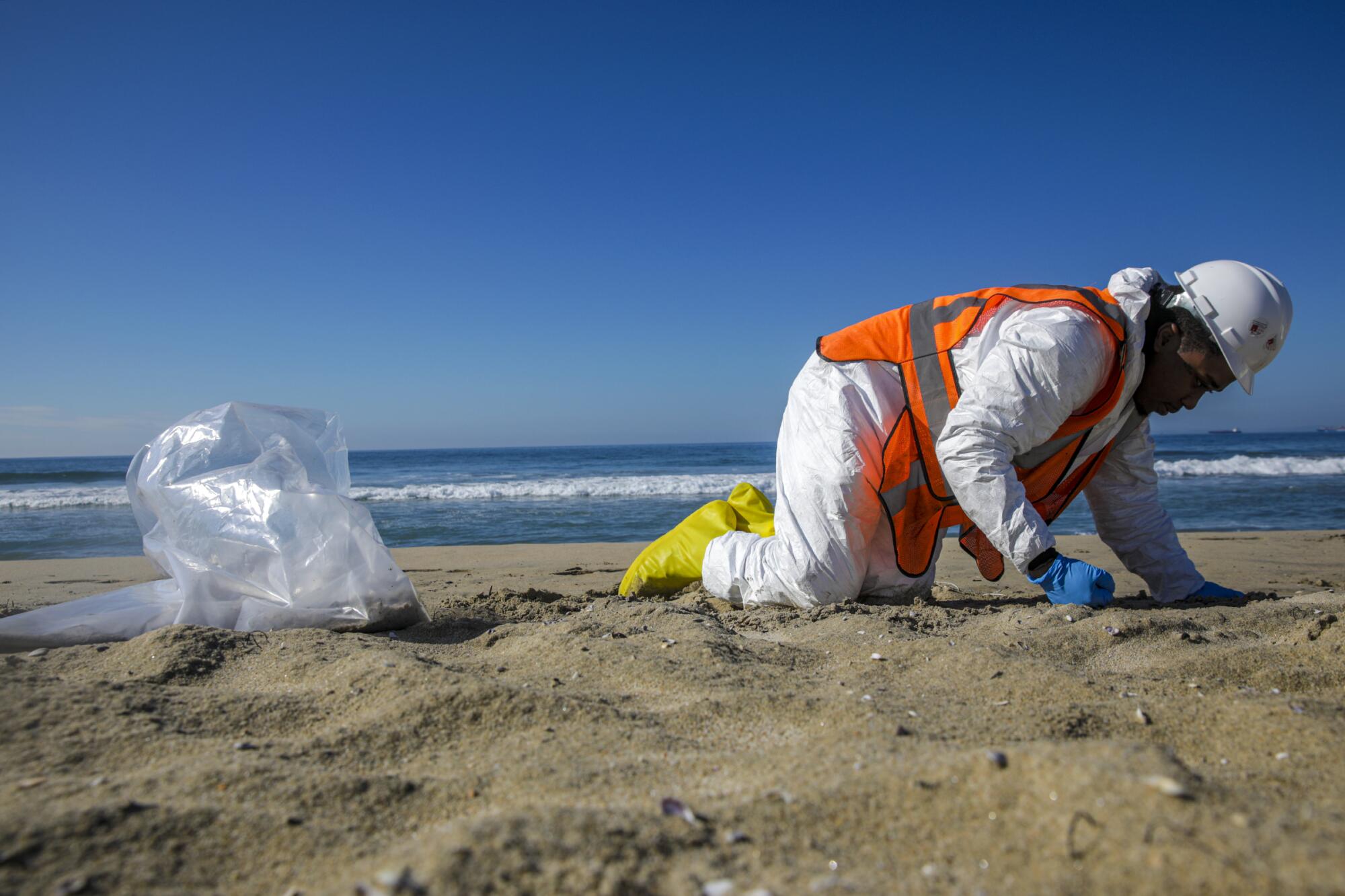 A man in a white hard hat and protective suit and blue gloves kneels while looking down on the sand