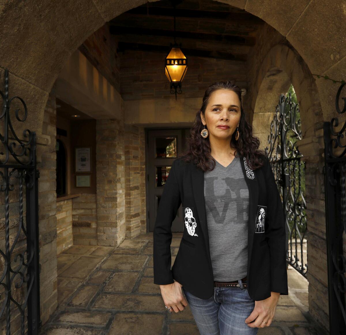 Larissa Fasthorse, wearing a grey t-shirt and a black blazer over jeans, is framed by a colonnade
