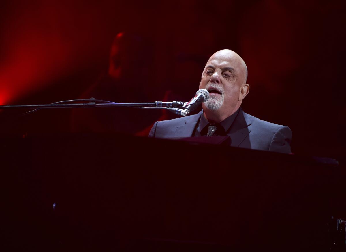 A bald man with a gray beard wearing a suit while singing into a microphone and playing piano