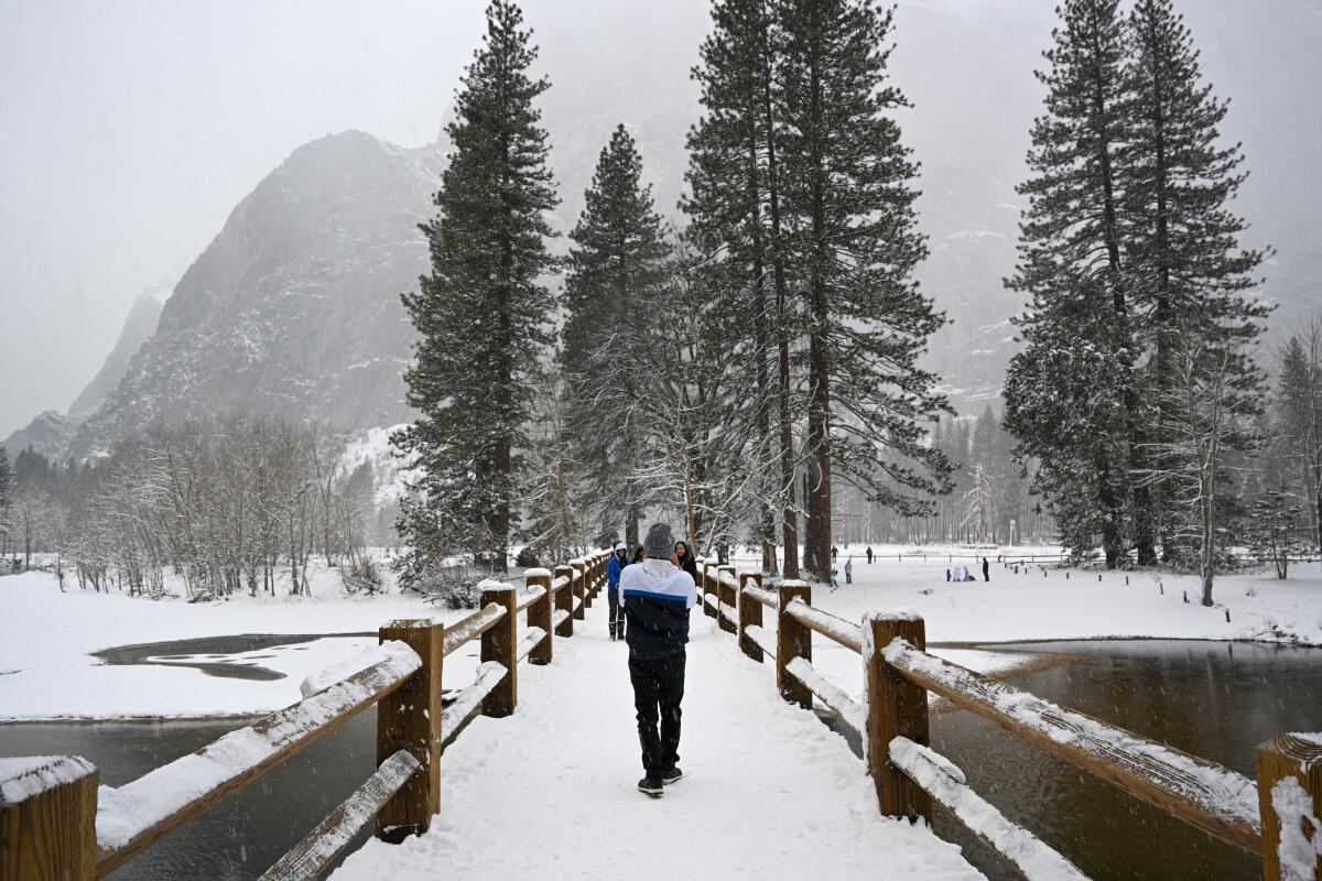 A person walks across a snow-covered bridge with mountains and trees in the background.