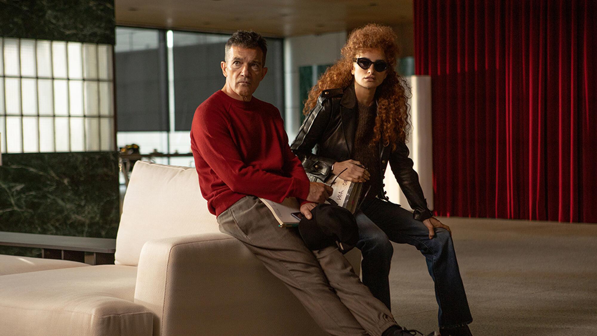 A man in a red sweatshirt and a woman with lots of long curly hair lean on the arm of a white sofa.