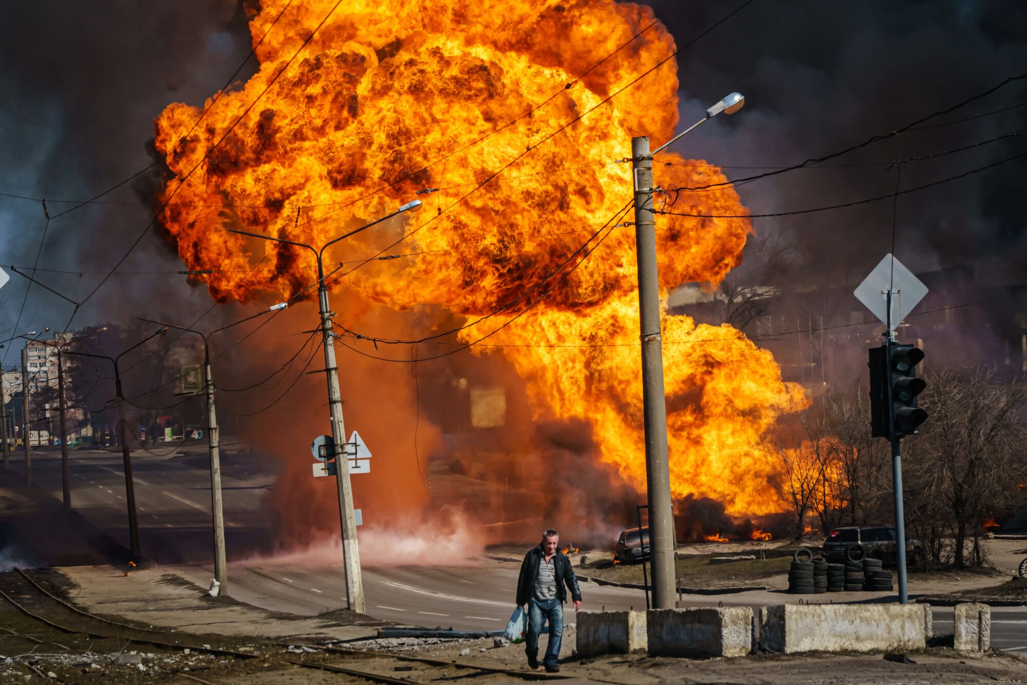 A small, lone figure of a man in the foreground walks away from a massive fiery explosion filling most of the background.