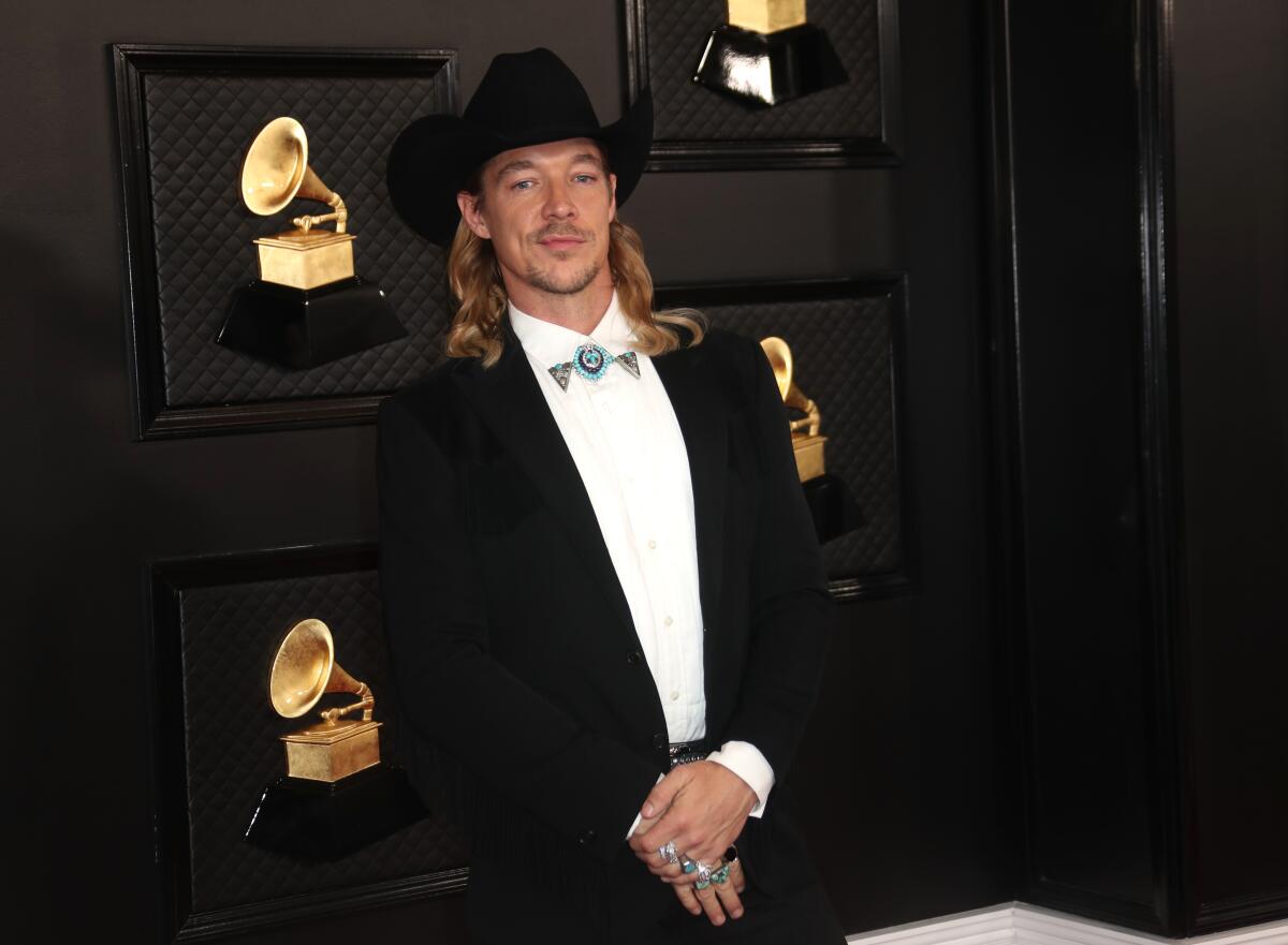 A man with long, blond hair posing in a black suit and black cowboy hat with Grammy awards behind him