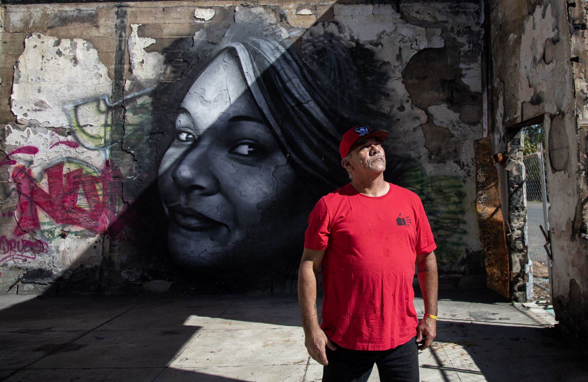 A man in a red T-shirt and cap stands in front of a mural of a woman