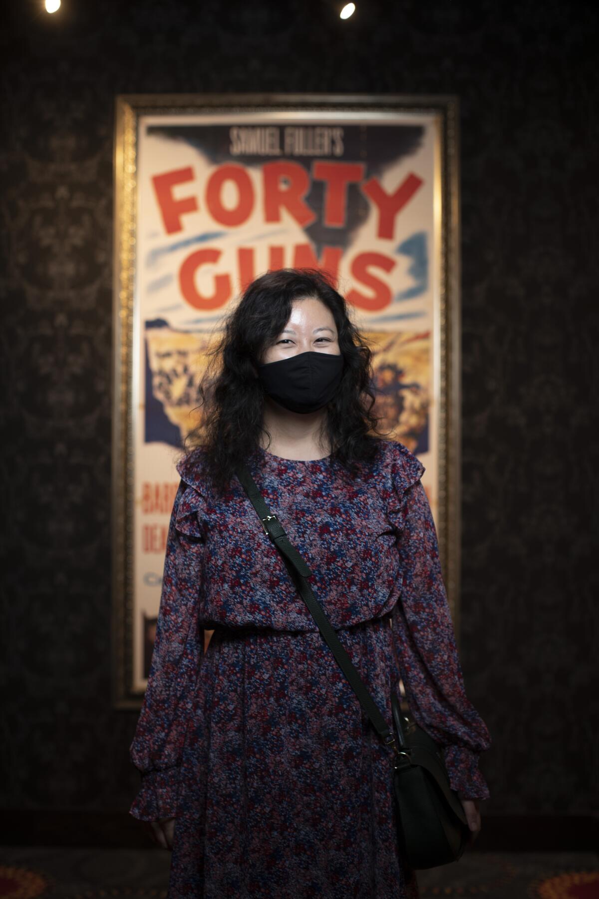 A woman in a mask and patterned dress stands in front of a "Forty Guns" movie poster