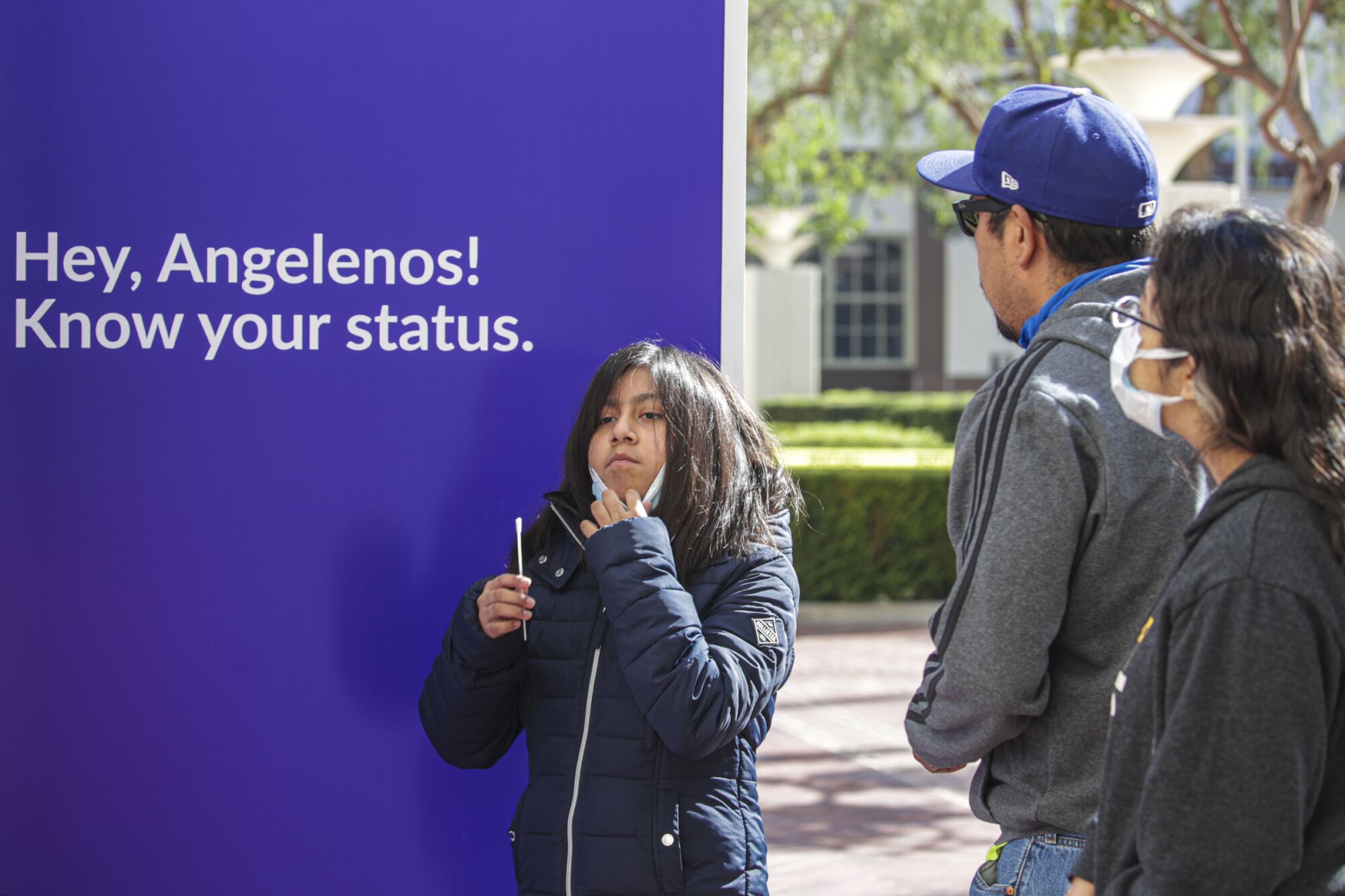 A girl, standing in front of a sign that says "Hey, Angelenos! Know your status," holds a test swab.