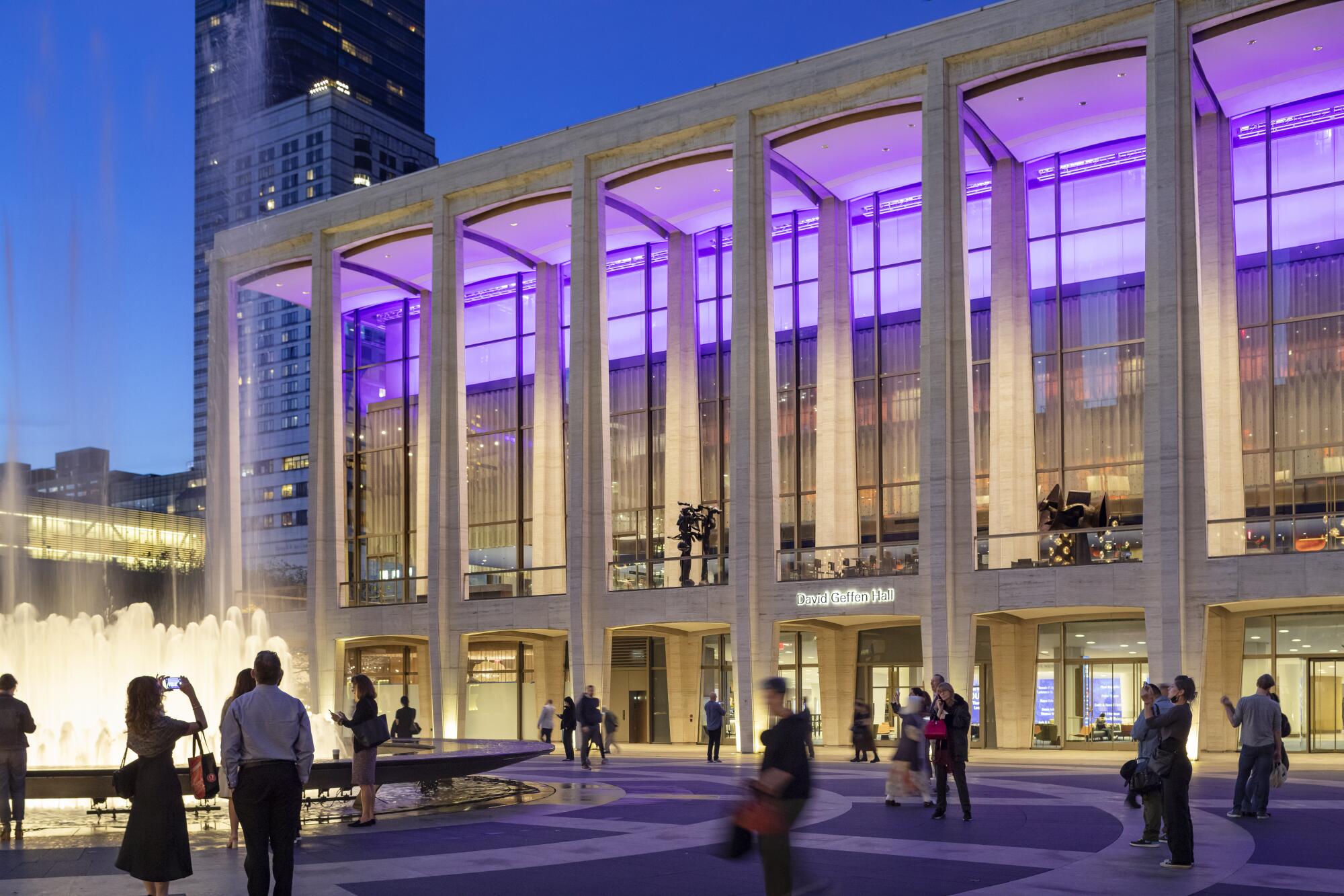 A Modernist buildling supported by gently tapering columns is illuminated with purple light from within.