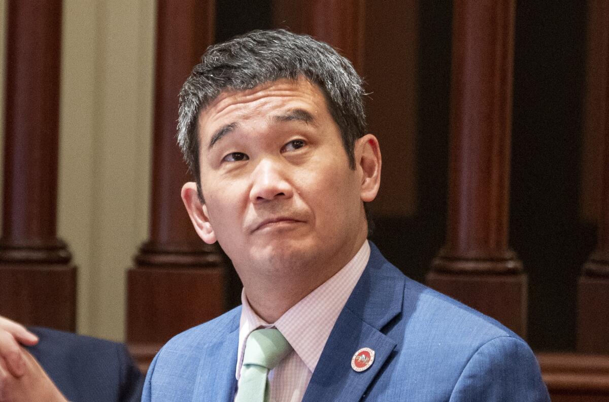 Dave Min, seen from the shoulders up in a blue suit jacket, looking up to his left against a backdrop of dark-wood columns