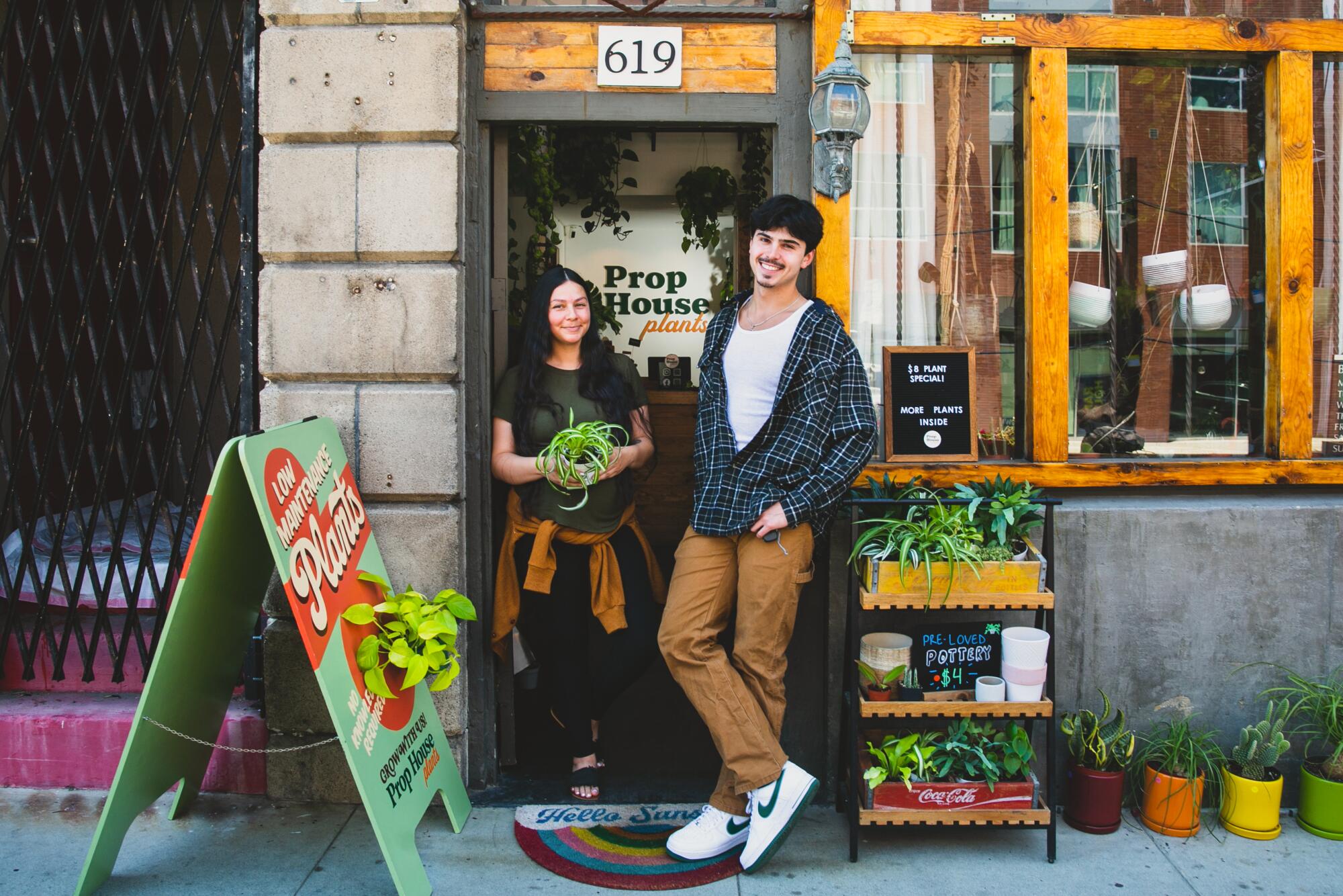 A young woman and a young man smile outside a door that reads "Prop House Plants."