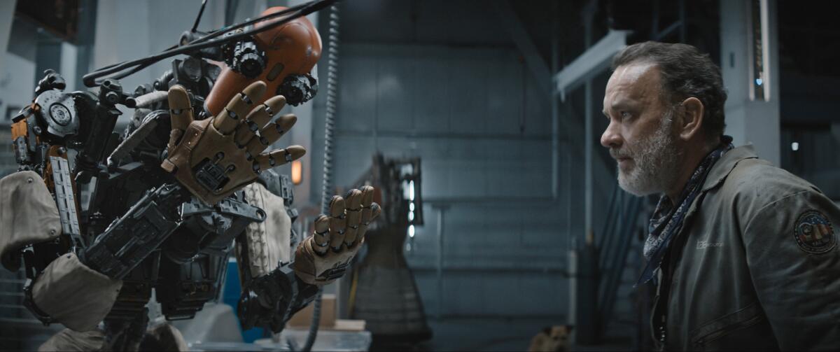 Tom Hanks stands opposite a robot that looks at its hands in wonder.