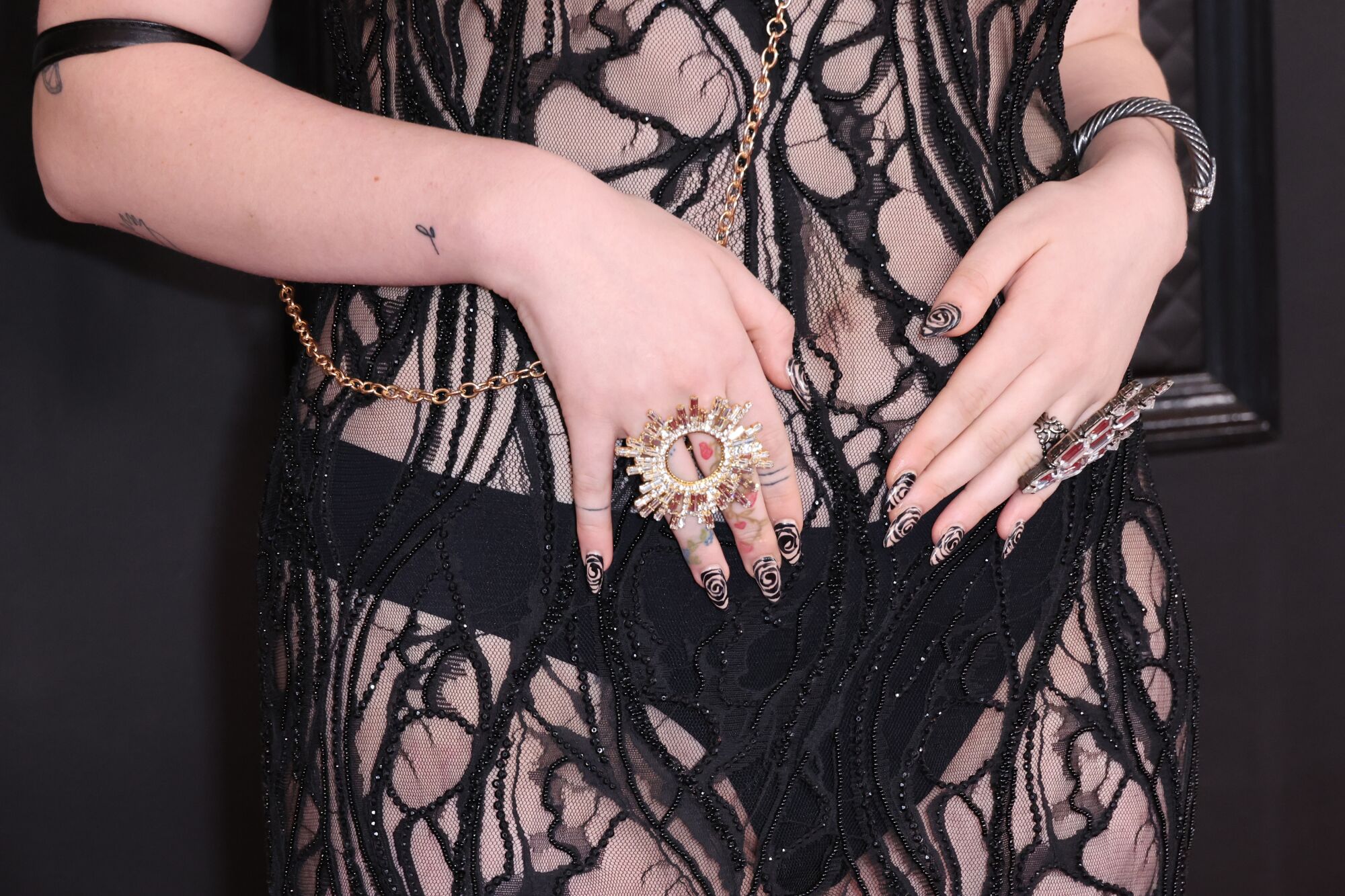 A torso covered in black and see through patterned mesh and hands with elaborate rings and fingernail polish.