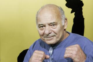 Burt Young wears a blue sweatshirt and holds up fighting fists.