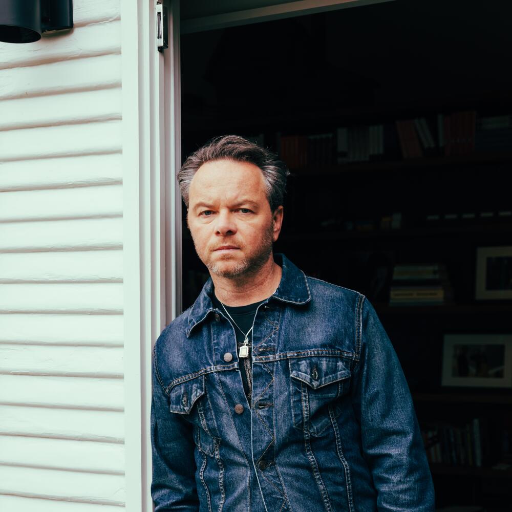 Noah Hawley, in a denim jacket and brown pants, stands in a doorway with his hands in his pockets.