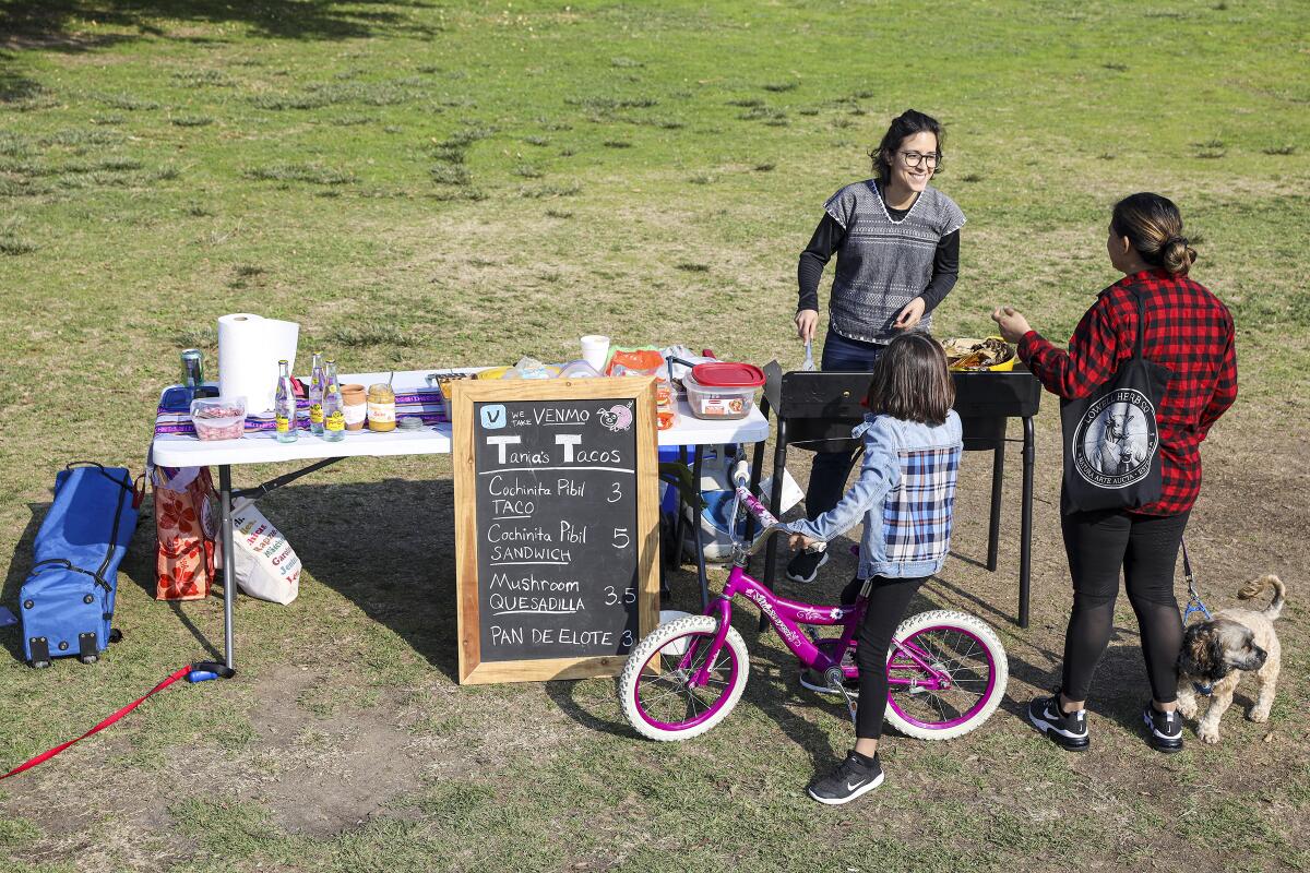 Tania Macin at her taco stand at Echo Park Lake with possible customers, a girl on a bike and a woman walking a dog.