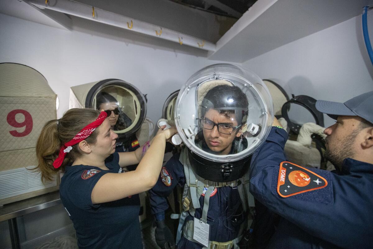 Mars Desert Research Station crew 212 members Zoe Townsend, left, and Vittorio Netti, right, attach a helmet to Camilo Zorro, before he and and others enter the airlock to go outdoors to collect soil samples. (Brian van der Brug / Los Angeles Times)