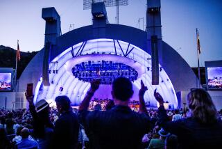 Silhouettes of concert attendees raising their hands in front of the Hollywood Bowl, which has a purple hue