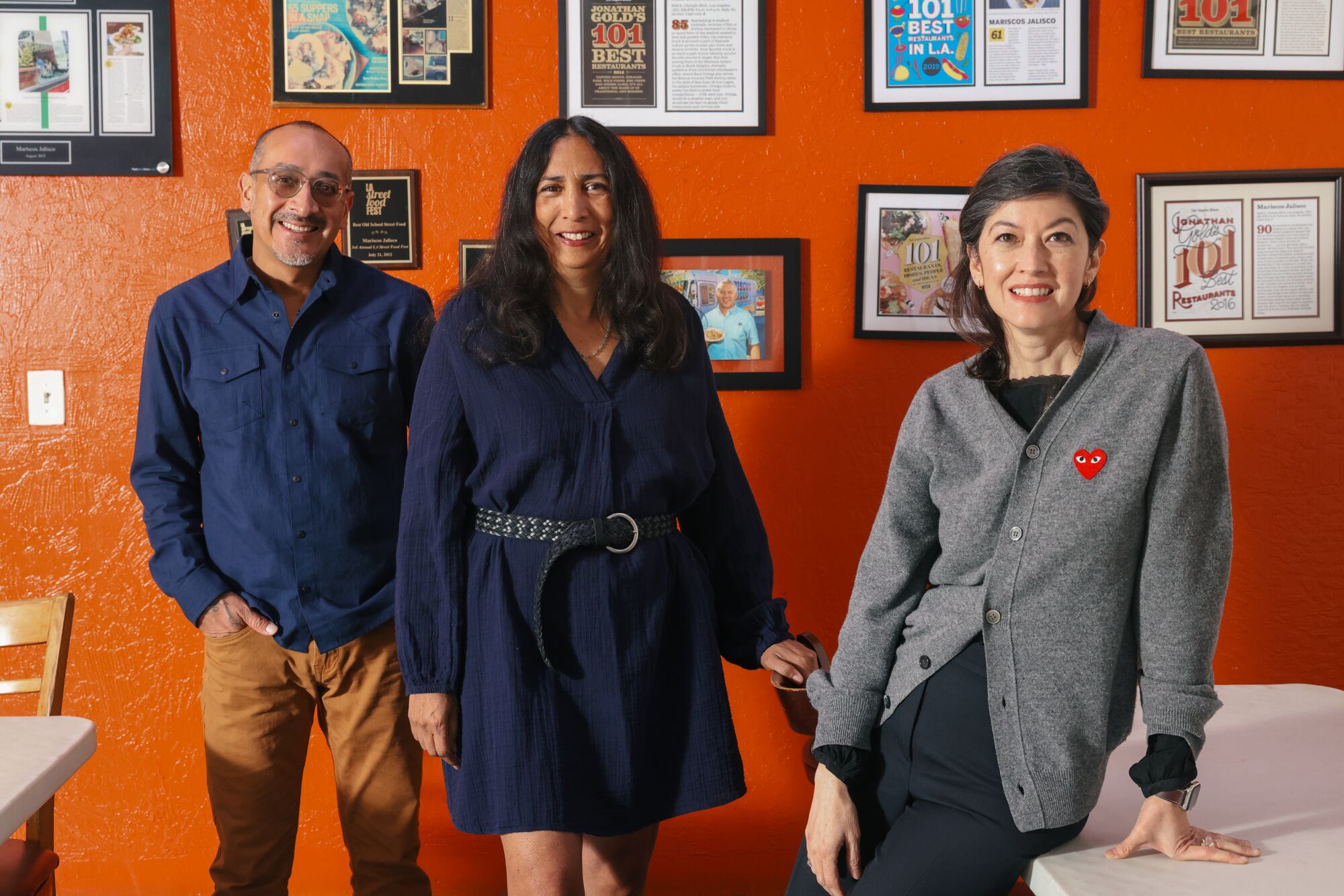 A man and two women stand before a bright orange wall that's decorated with framed restaurant reviews