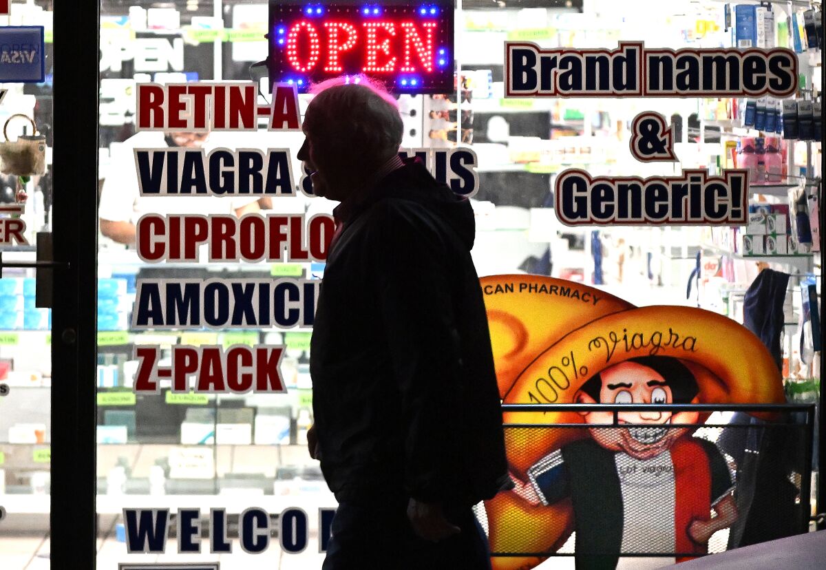 A man with a hooded jacket in silhouette near a pharmacy's glass window stenciled with drug ads