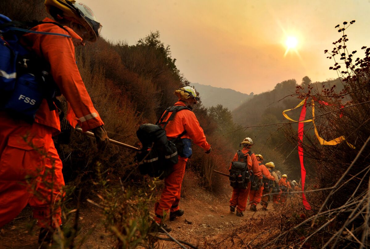 Women wearing orange firefighting suits walk along a dirt trail in hazy air 