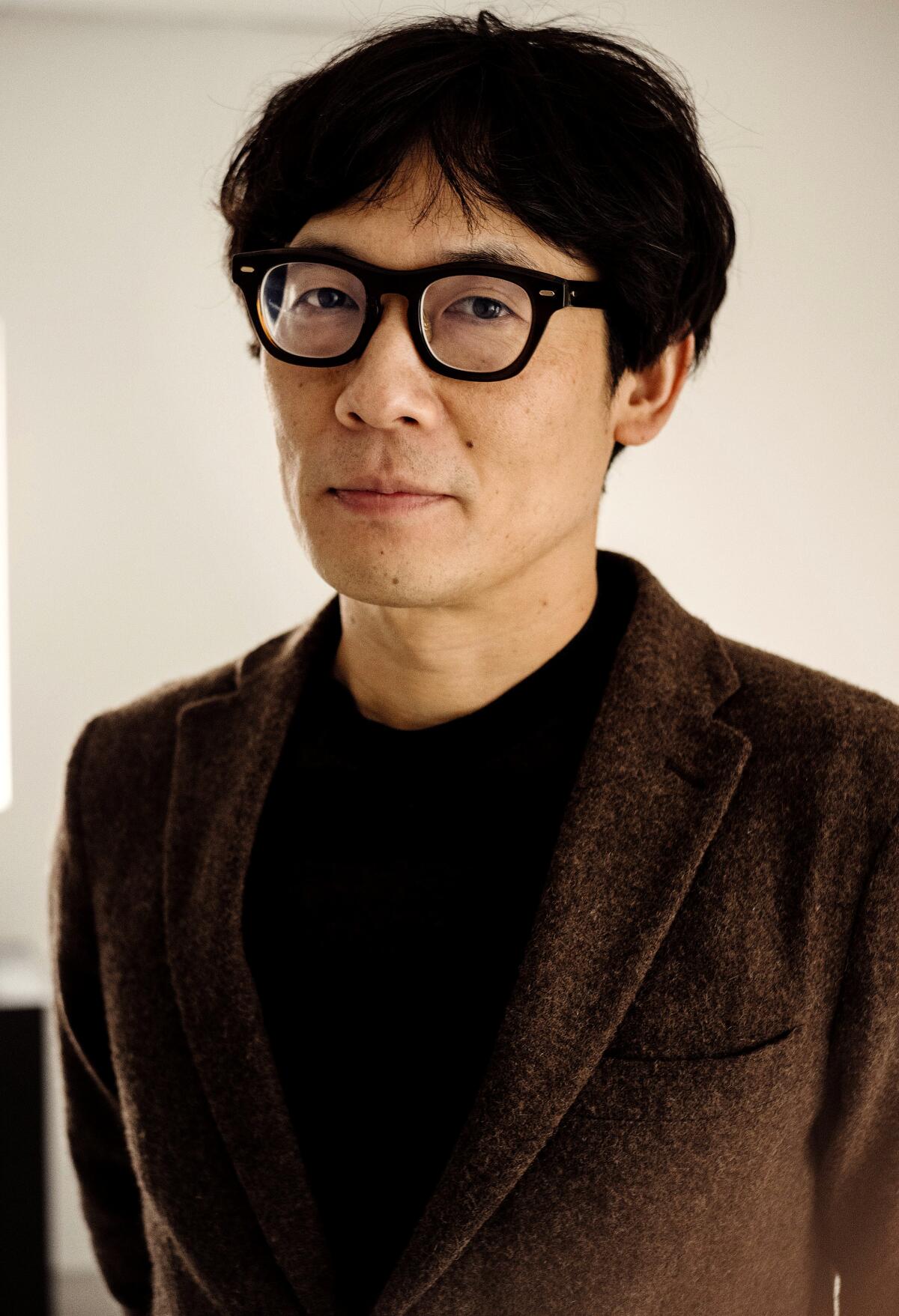 A  man with dark hair and square glasses in a dark shirt and jacket.